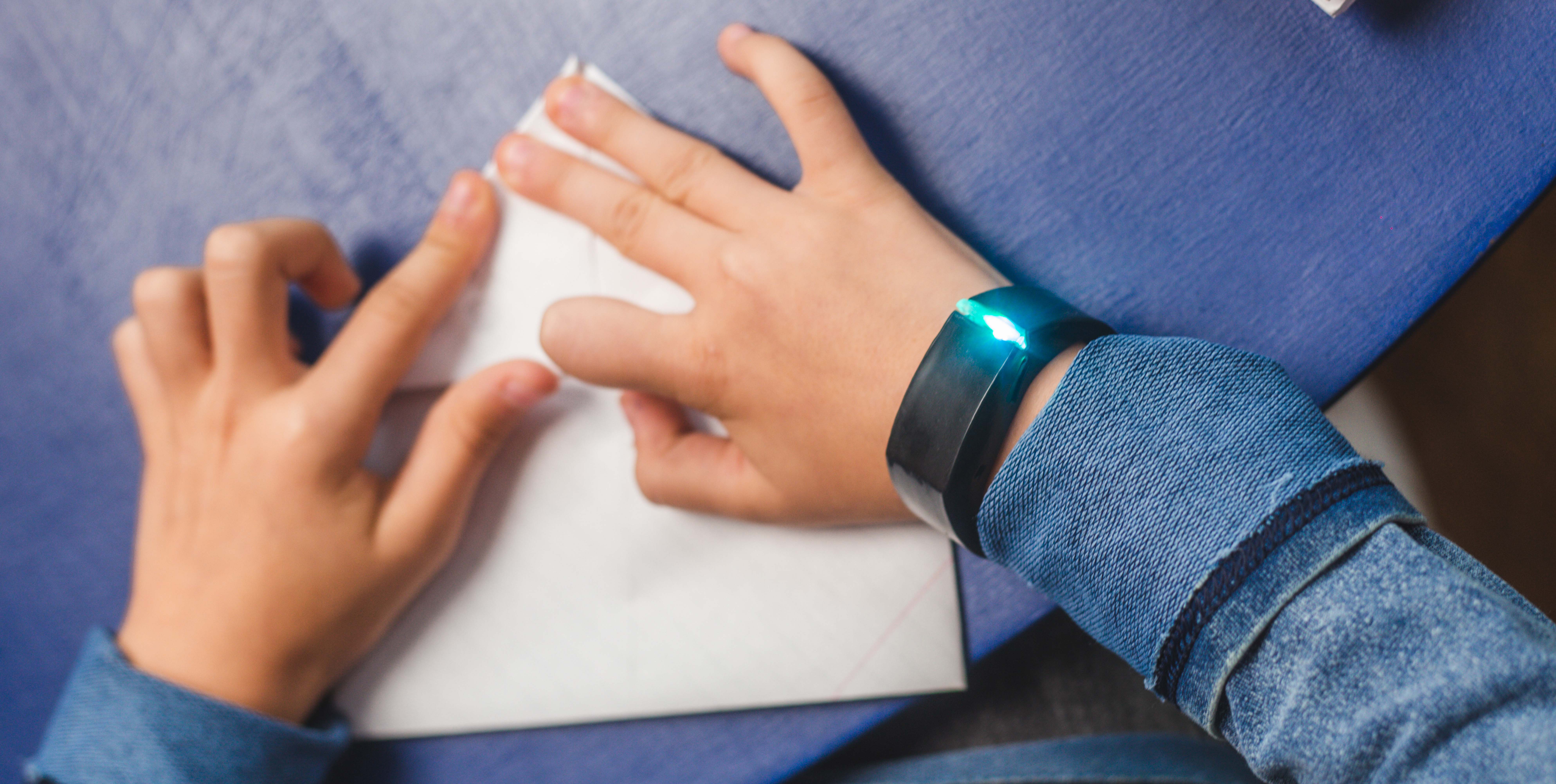 Child wearing a Tully smart bracelet with a glowing LED light while folding a paper, demonstrating the bracelet's discreet design and real-time emotional tracking feature.